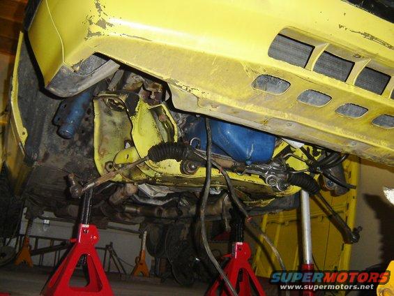 Yellow car underside, steering rack visible. Supported by jack stands