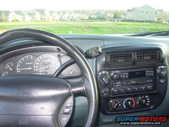 Dashboard view from driver's seat, showing steering wheel, gauges, radio, and air vents. Houses visible outside