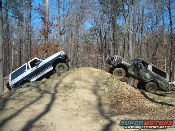 Two Ford Broncos climbing a dirt hill off-road. One is white and blue, the other is camouflaged