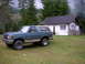 1990 Ford Bronco - A 1990 Ford Bronco sits in a grassy area near a rustic building with a wooden frame and a shingled roof.