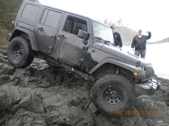 Dark gray Jeep Wrangler four-door on a rocky shore, two people standing nearby