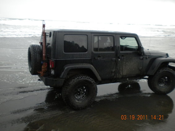 Black four-door Jeep Wrangler parked on wet sand near ocean waves. Spare tire mounted on rear with a Hi-Lift jack attached
