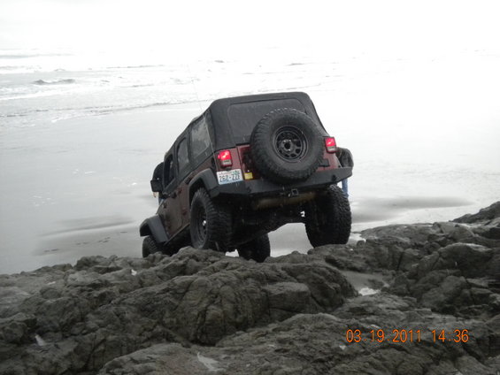 Jeep Wrangler with soft top on rocky beach, rear view. License plate reads 262-ZZZ