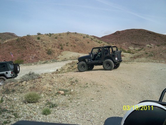 Black Jeep Wrangler driving on a dirt road in a desert landscape. Another Jeep is visible to the left