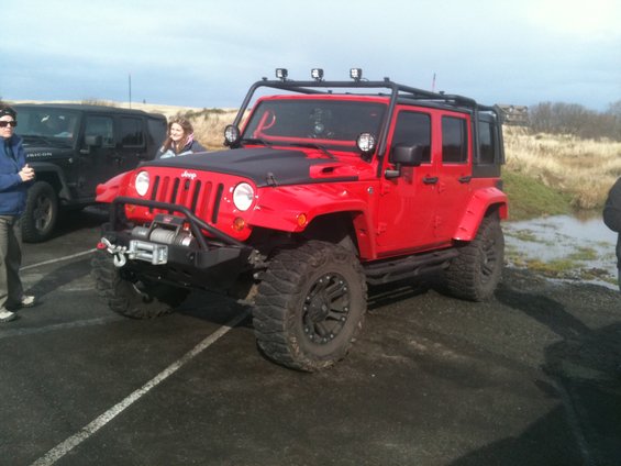 Red four-door Jeep Wrangler with a black hood and roof rack, parked on asphalt