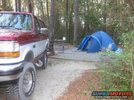 Red and silver 1995 Ford Bronco parked at a campsite with a blue tent and chairs