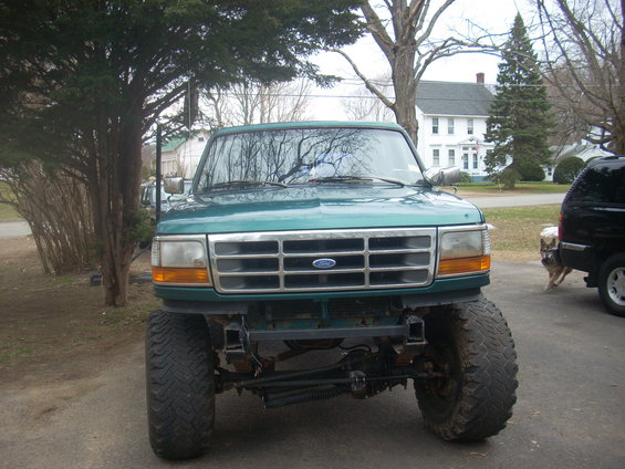 Green Ford F-Series truck with large tires, front view. Another black SUV is parked to the right