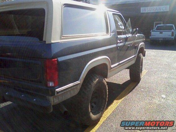 Dark blue Ford Bronco with a white top, parked in a lot. Another white truck is in the background. Sign says 'INSURANCE'