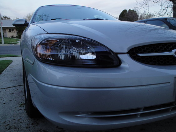 Silver sedan front end, close-up, showing headlights and grille. Another dark vehicle is partially visible behind it