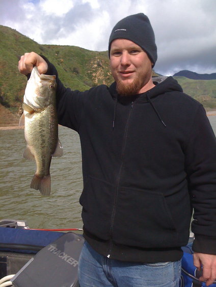 Man in black hoodie and beanie holds a large bass while standing on a boat with 'TRACKER' visible on a seat