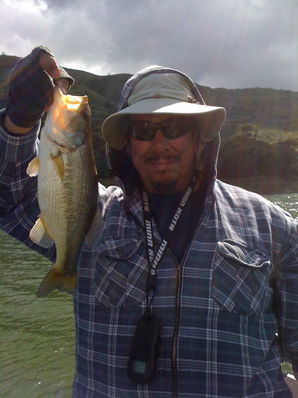 Guy in a hat and sunglasses holds up a bass he caught while fishing on a lake