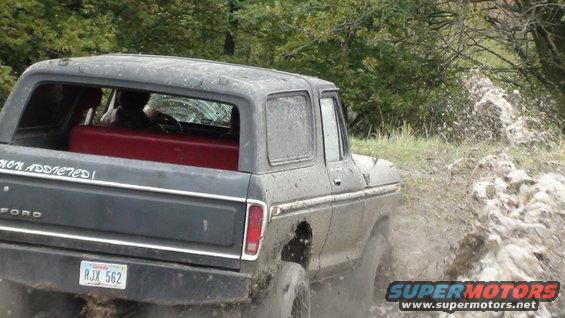 Dark gray 1978 Ford Bronco driving through mud, text on rear window reads 'Addicted!'
