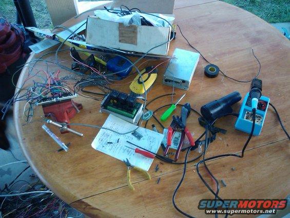 Workbench with a red vise holding a circuit board, wires, soldering iron, pliers, and other electronic components