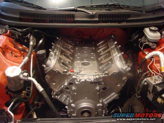 Engine bay with a V8 engine installed, orange fender, brake fluid reservoir, and Supermotors.net watermark