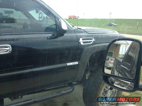 Black pickup truck side view, showing 'EL JEFE' badge and chrome trim. Side mirror reflects other vehicles