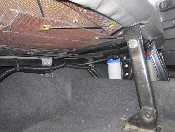 Underneath a car seat, black carpet, wiring, and two white canisters with blue tops