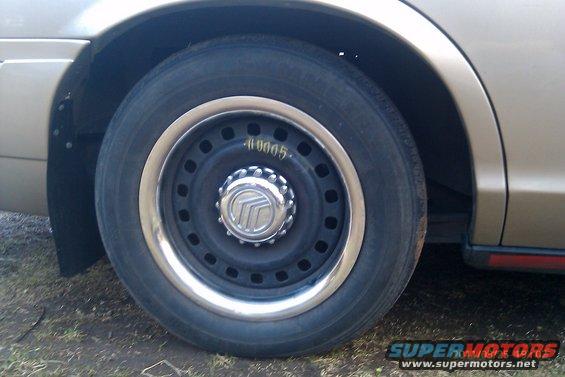 Gold car wheel with a chrome trim ring and a Mercury logo center cap. The tire has visible text