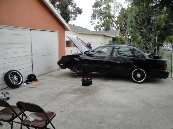 Black sedan parked in driveway with hood and trunk open, front wheel removed. Tools and spare tire nearby
