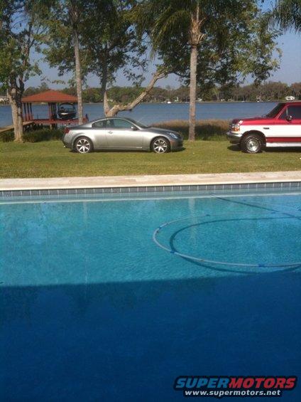 2007 Infiniti G35 coupe parked on grass next to a red and white pickup truck, with a pool in the foreground and a lake in the background