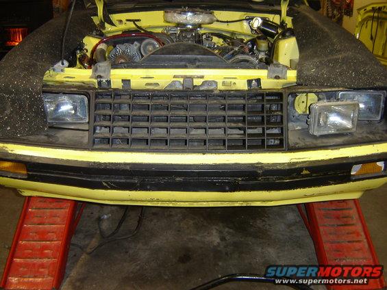 Yellow 1979 Ford Mustang front end with hood open, showing engine and grille. Car is on red ramps