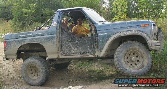 Dark green Ford Bronco with two people inside, off-roading. Website URL visible on the side