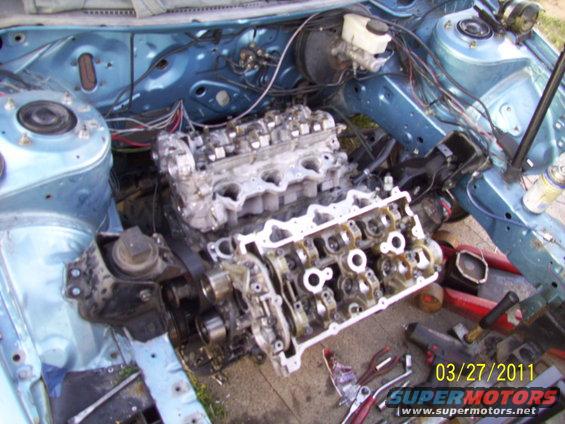 engine bay with engine partially disassembled, showing engine block and cylinder head removed. Tools and parts are scattered around