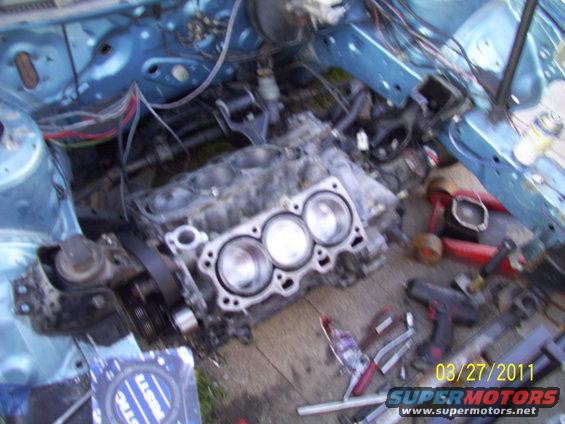 Engine block with three cylinders visible, disassembled engine parts, tools, and blue car frame
