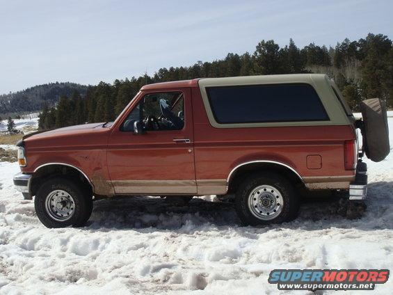Side view of a red and tan Ford Bronco parked in the snow with trees in the background