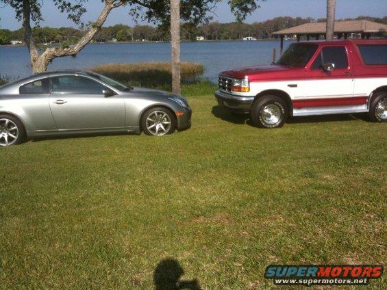 Silver Infiniti coupe and red/white Ford Bronco parked on grass near a lake