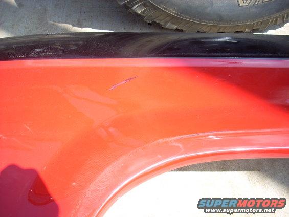 Close-up of a red vehicle fender with a scratch and a black trim piece above it. A tire is visible at the top