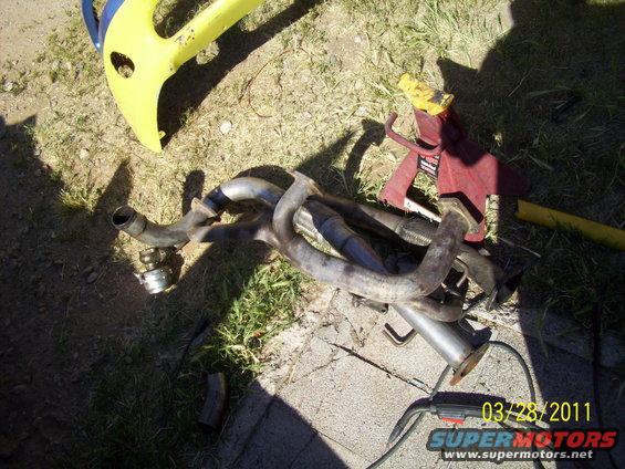 Exhaust manifold and pipes on the ground next to a yellow plastic object and a red jack stand