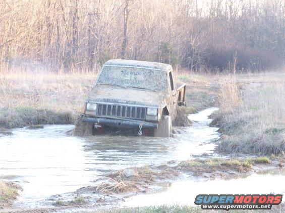 Muddy Jeep Cherokee stuck in a water-filled ditch, trees in background
