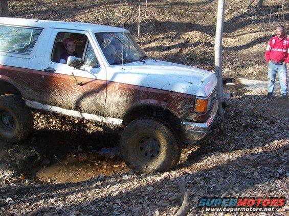 Mud-splattered white and brown Ford Bronco driving through a muddy wooded area