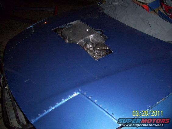 Blue car hood with a cutout revealing engine components, riveted seams visible