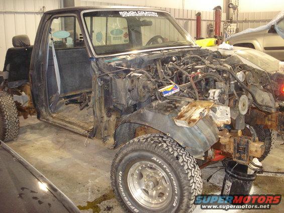 Disassembled truck in a garage, engine bay empty, doors open. Rough Country sticker on windshield