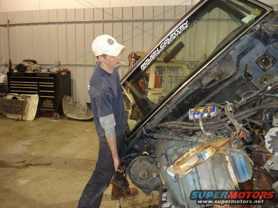 Man in blue shirt and white cap working on a disassembled blue truck engine bay, with "ROUGH COUNTRY" sticker on windshield