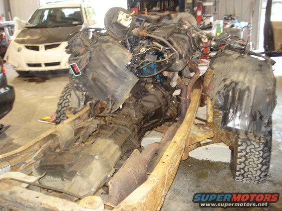 Engine and transmission assembly mounted on a rusty frame, with a white car in the background