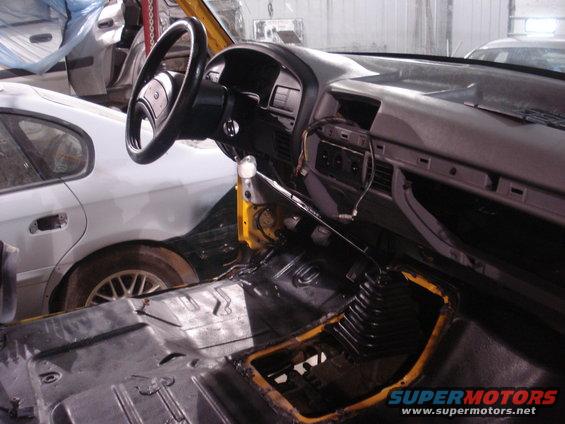 Interior of a 1993 Ford F-150 with dashboard and center console removed, showing bare metal floor and a white car in the background