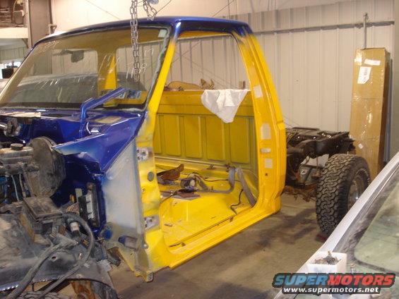 Truck cab, stripped down and painted blue and yellow, suspended by chains in a workshop