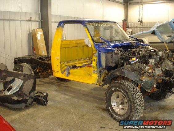 Pickup truck cab and chassis disassembled in a garage, yellow and blue paint, engine exposed