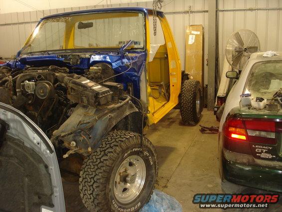 Blue and yellow Ford F-150 truck body with engine exposed, undergoing restoration. A green sedan is parked nearby
