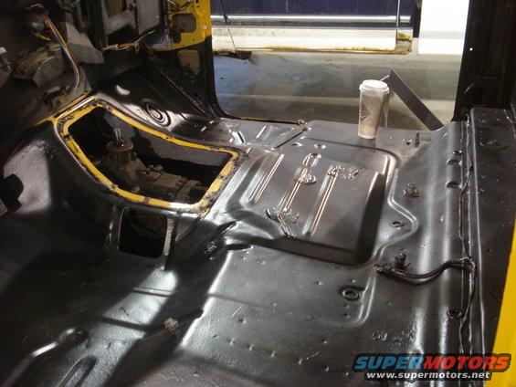 Vehicle cab interior with exposed floor pan and transmission tunnel, yellow paint visible on dashboard. Coffee cup on floor