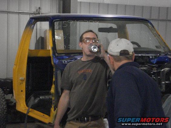 Two guys in a garage, one drinking from a can, next to a yellow and blue truck cab being worked on