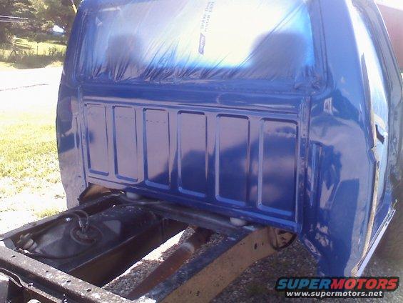 Rear view of a blue pickup truck bed with plastic sheeting covering the rear window area. The frame and fuel tank are visible