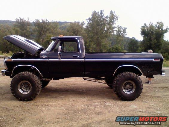 Side view of a black lifted pickup truck with the hood open. It has large off-road tires and chrome trim