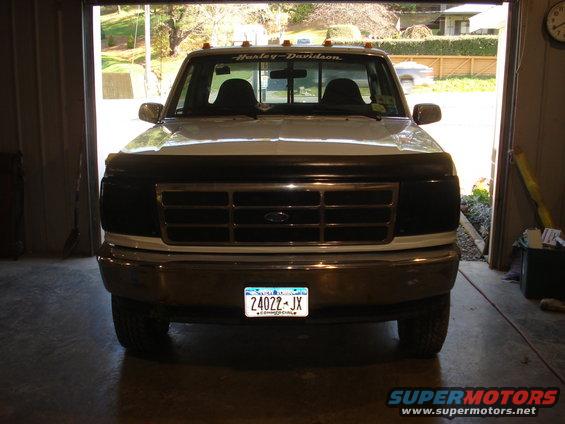 Front view of a white Ford F-150 truck with a black grille guard and Harley-Davidson banner on the windshield. License plate reads 24022-JX