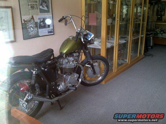 Old green motorcycle with custom handlebars and seat, parked indoors near display cases