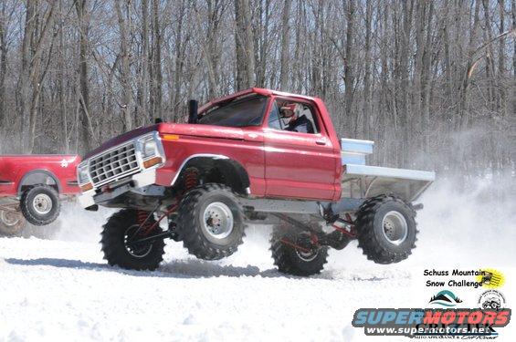 red lifted pickup truck jumping in snow, another red truck behind it