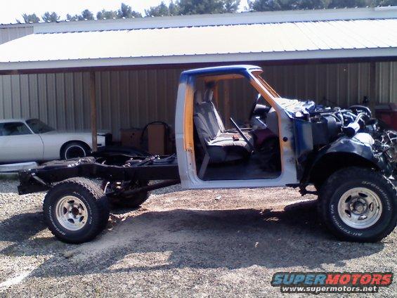 Truck chassis with cab, engine removed, parked outside a building. Another car is visible in the background