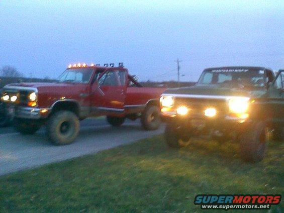 Two lifted trucks, one red and one dark colored, parked outside at dusk with their headlights on